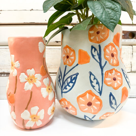 The Brightside Store | Juniper Vase Peach Two hand-painted ceramic planters with a floral pattern, one in terracotta pink and one in off-white, shown with green leaves on a white wooden surface.