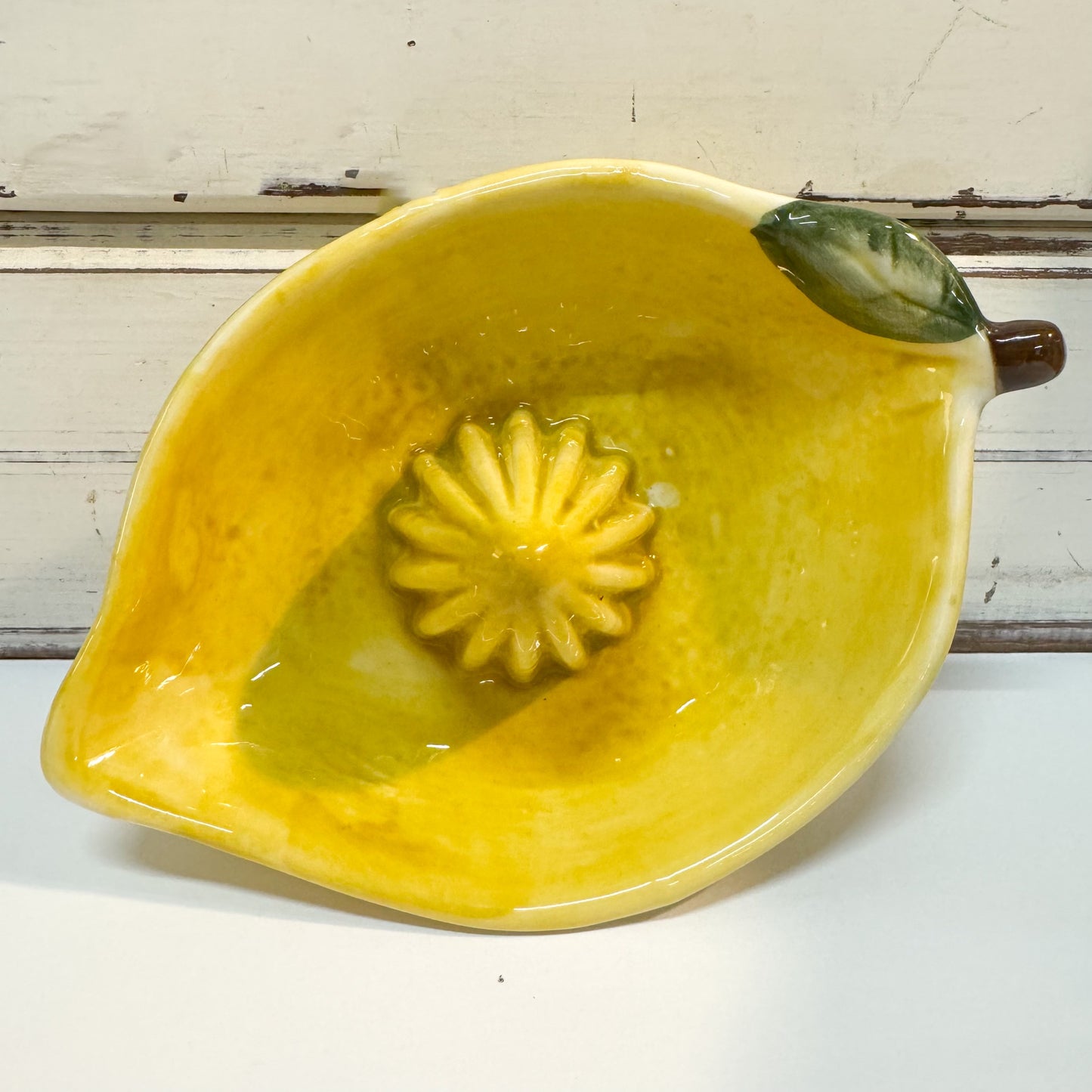 The Brightside Store | Limone Lemon Juicer A yellow ceramic lemon-shaped bowl or dish with a molded citrus pulp texture in the center and a green leaf detail on the rim, shown on a white surface against a rustic white wooden background.