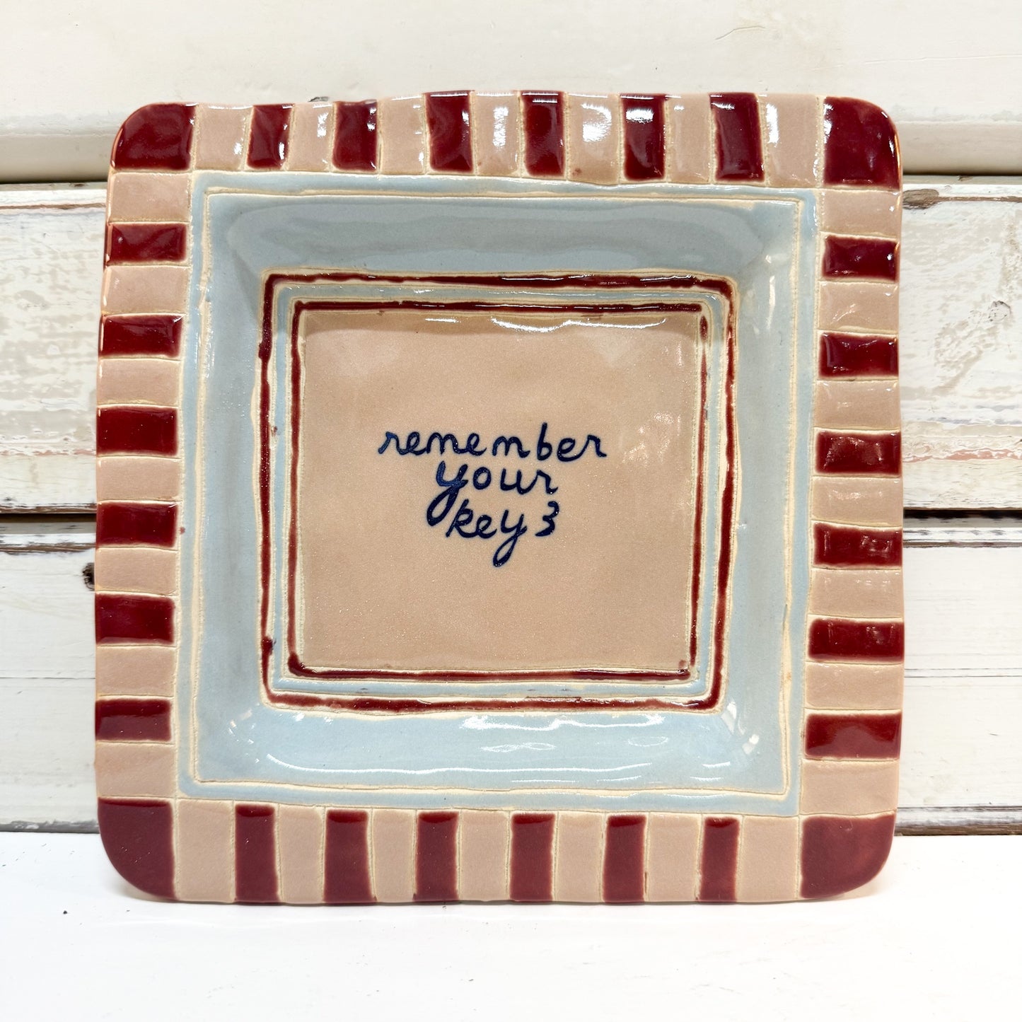 The Brightside Store | Lingo Tray - Keys
A square ceramic dish with a striped border in beige and dark red, featuring a light blue inner border and a beige center with the handwritten phrase 'remember your key 3' in dark blue ink.