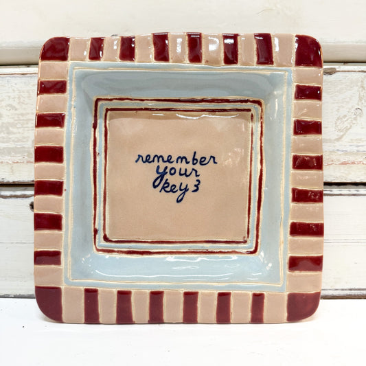The Brightside Store | Lingo Tray - Keys
A square ceramic dish with a striped border in beige and dark red, featuring a light blue inner border and a beige center with the handwritten phrase 'remember your key 3' in dark blue ink.