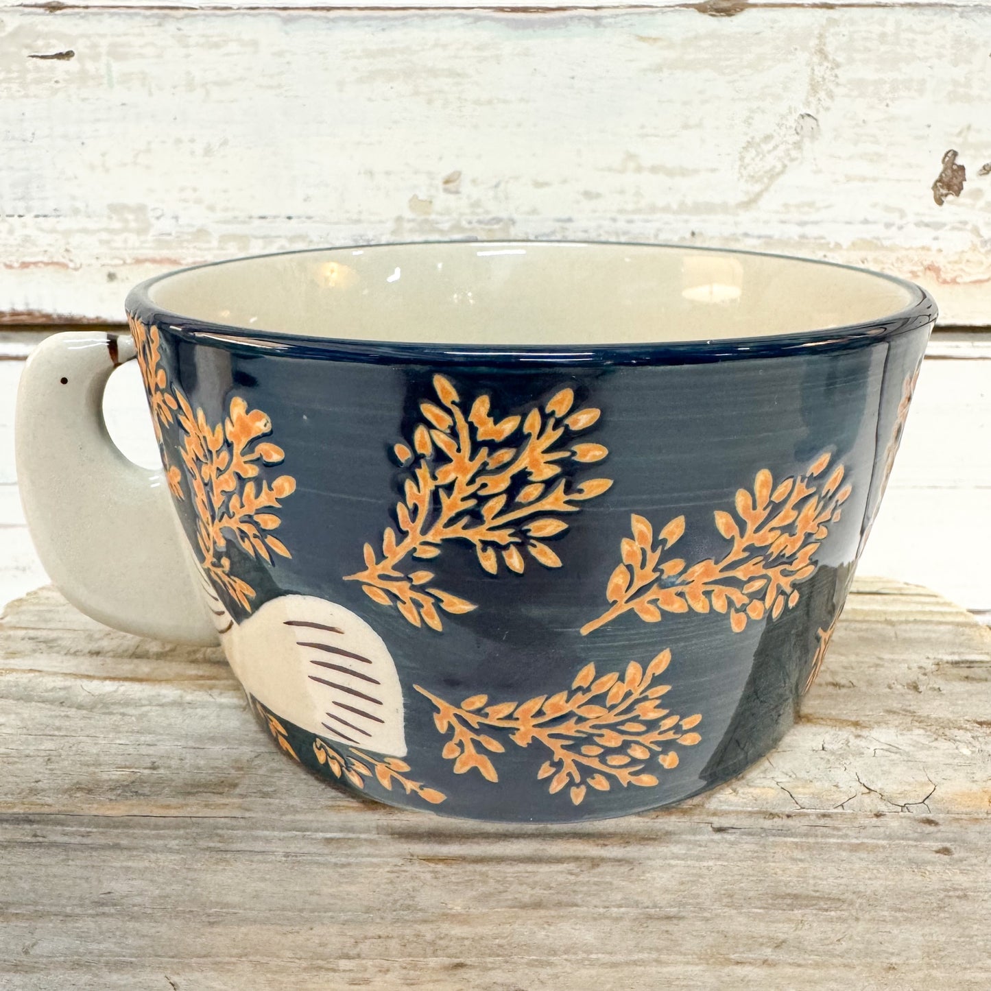 A ceramic bowl with a dark navy blue exterior decorated with gold botanical leaf patterns and a cream-colored handle and interior, set on a rustic wooden surface.