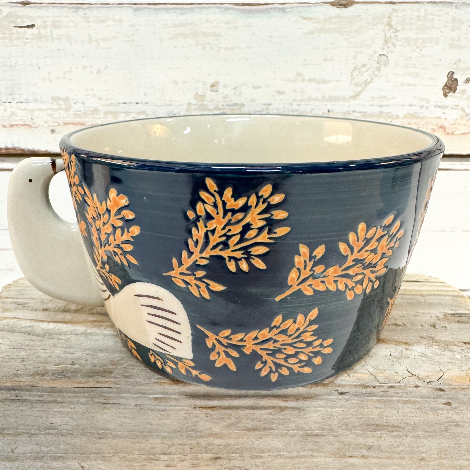 A ceramic bowl with a dark navy blue exterior decorated with gold botanical leaf patterns and a cream-colored handle and interior, set on a rustic wooden surface.