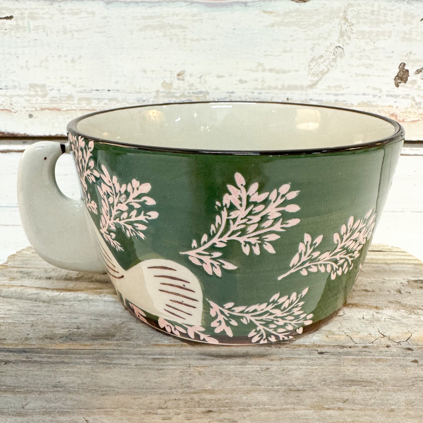 A ceramic mug with a dark green exterior decorated with light pink botanical leaf patterns and a white bird motif, set on a rustic wooden surface against a whitewashed wood background.