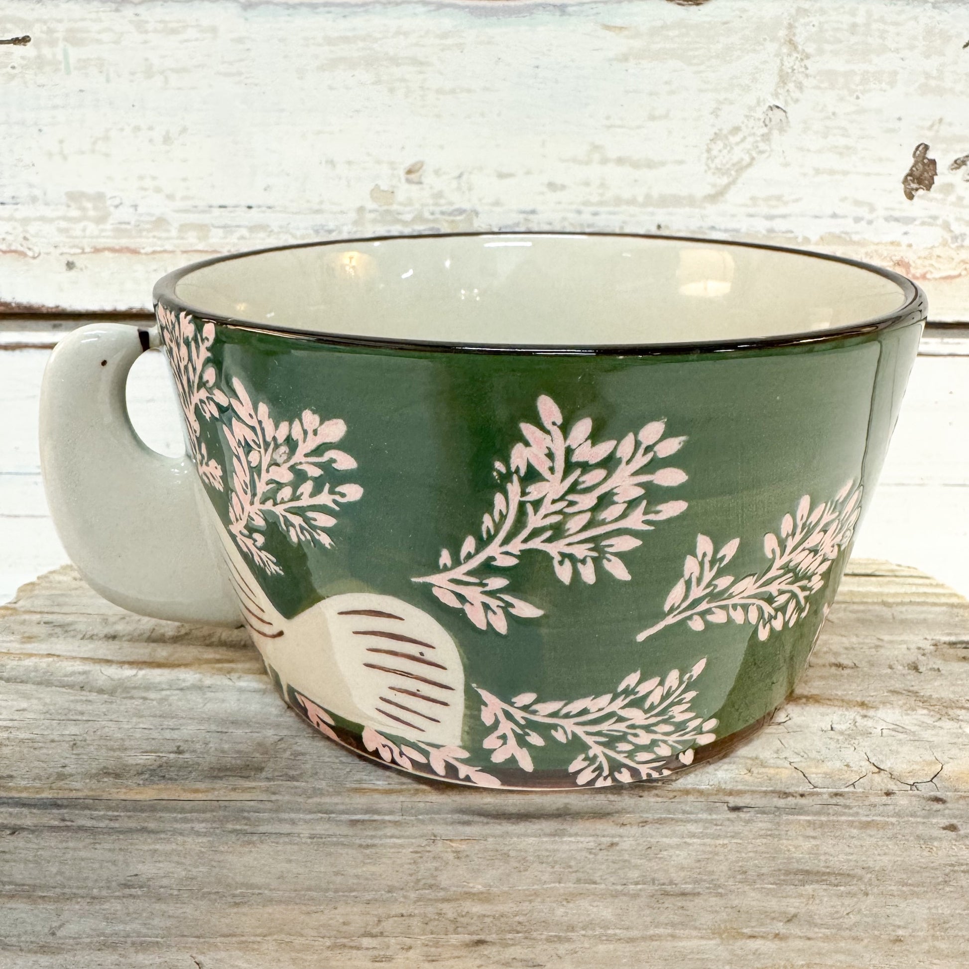 A ceramic mug with a dark green exterior decorated with light pink botanical leaf patterns and a white bird motif, set on a rustic wooden surface against a whitewashed wood background.