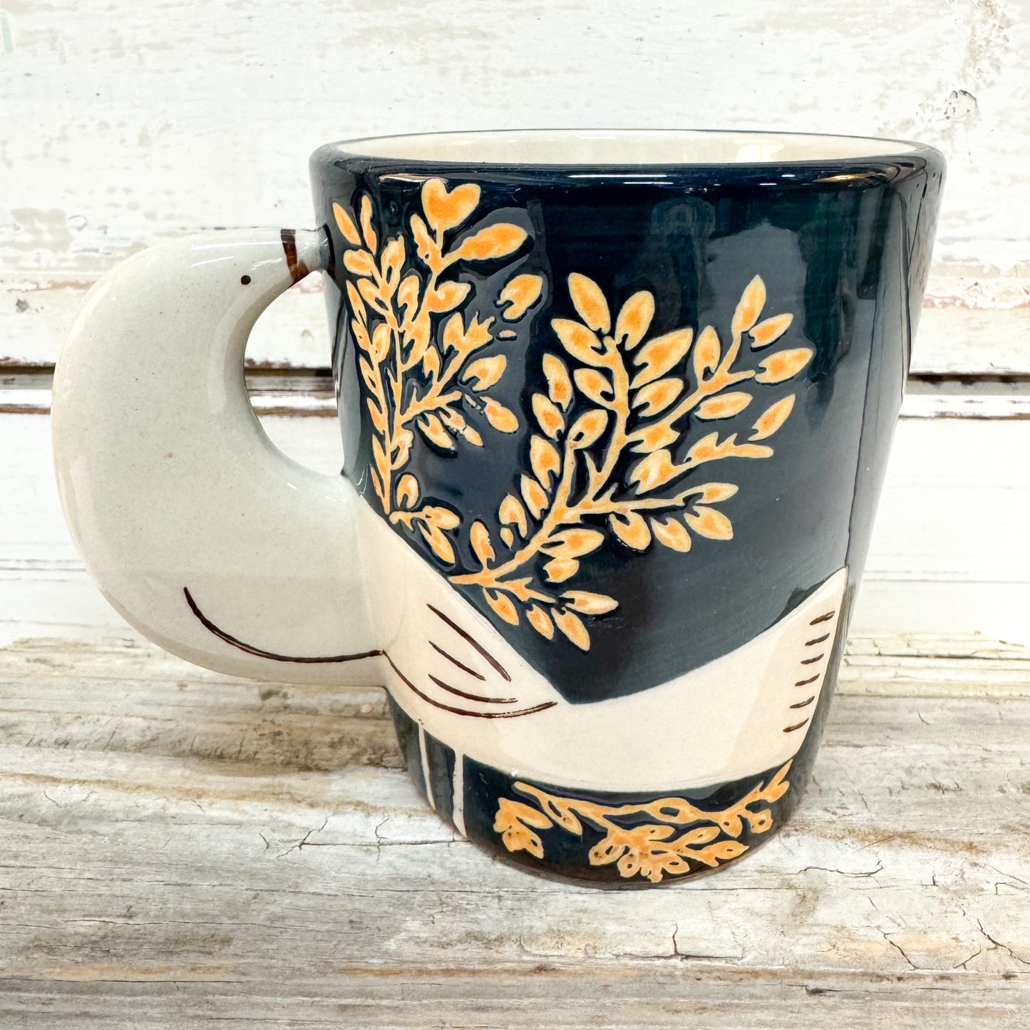 A ceramic coffee mug with a dark navy blue body, decorated with gold leaf patterns and a cream-colored handle and lower section, set against a rustic white wood background.