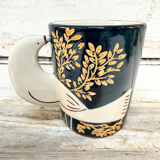 A ceramic coffee mug with a dark navy blue body, decorated with gold leaf patterns and a cream-colored handle and lower section, set against a rustic white wood background.
