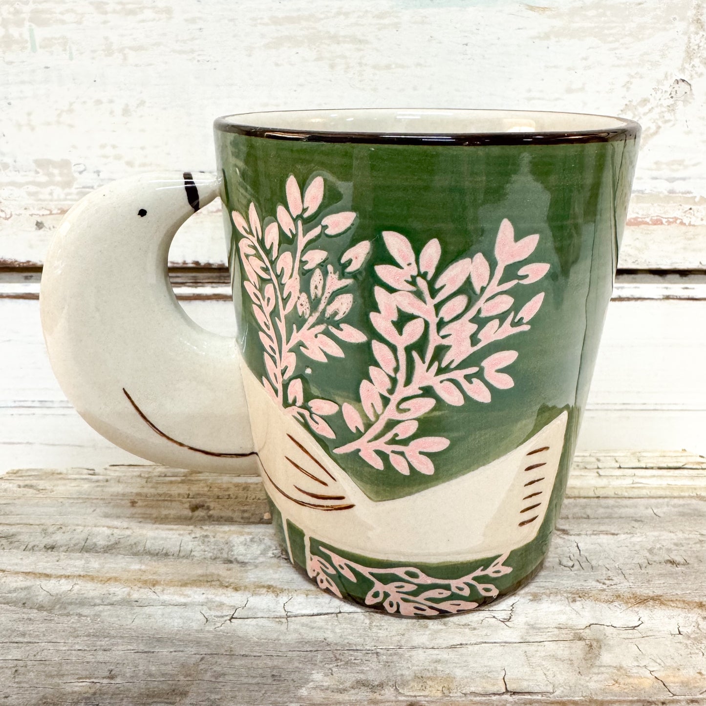A ceramic mug with a bird-shaped handle, featuring a dark green body decorated with pink leafy branches and cream-colored bird details, set against a rustic white-washed wood background.