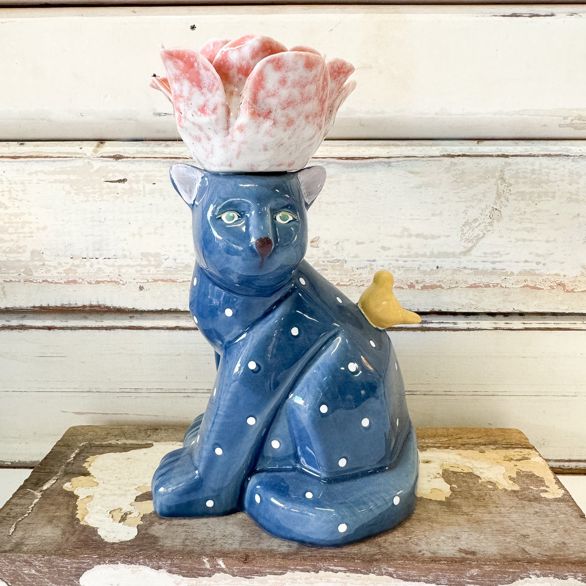 A blue ceramic cat candleholder with white polka dots, a pink and white flower on its head, and a small yellow bird on its back, displayed on a rustic wooden surface against a white distressed wood plank background.