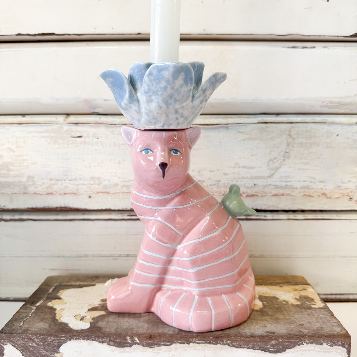 A pink ceramic cat candle holder with white stripes, a blue-gray flower-shaped top, and a small green bird detail, shown holding a white candle against a rustic white wood background.