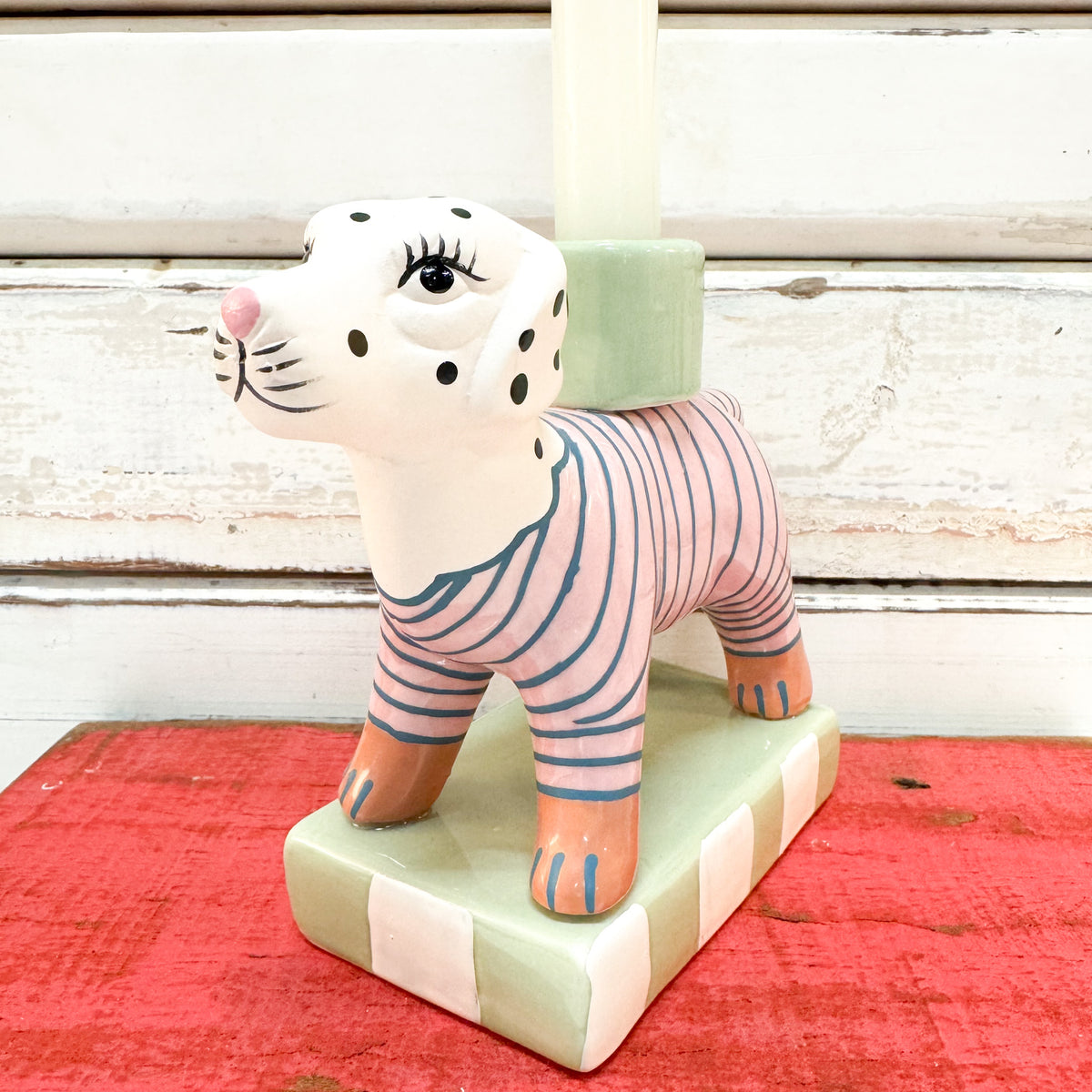 A ceramic cat-shaped candle holder with a white body, black spots, pink and blue stripes, and peach paws, holding a white candle on a striped mint-green and white base against a rustic white wood background and red surface.