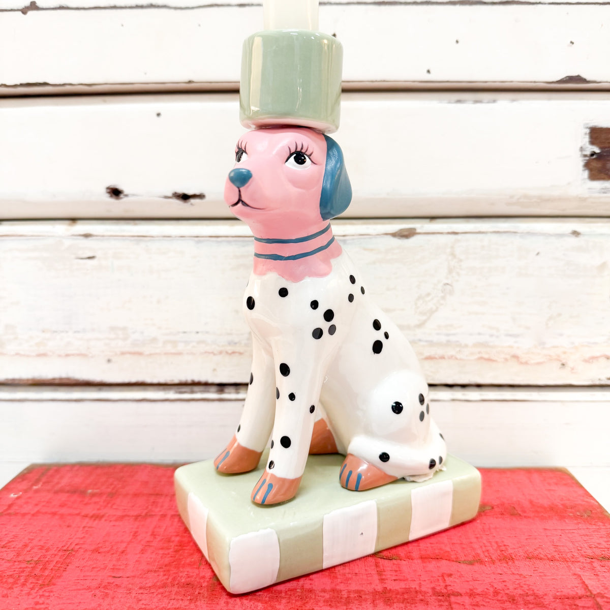 A ceramic dalmatian dog figurine candleholder with a pink face and ears, black spots, a blue nose, and a mint green candle holder on its head, sitting on a striped base against a rustic white wood background.