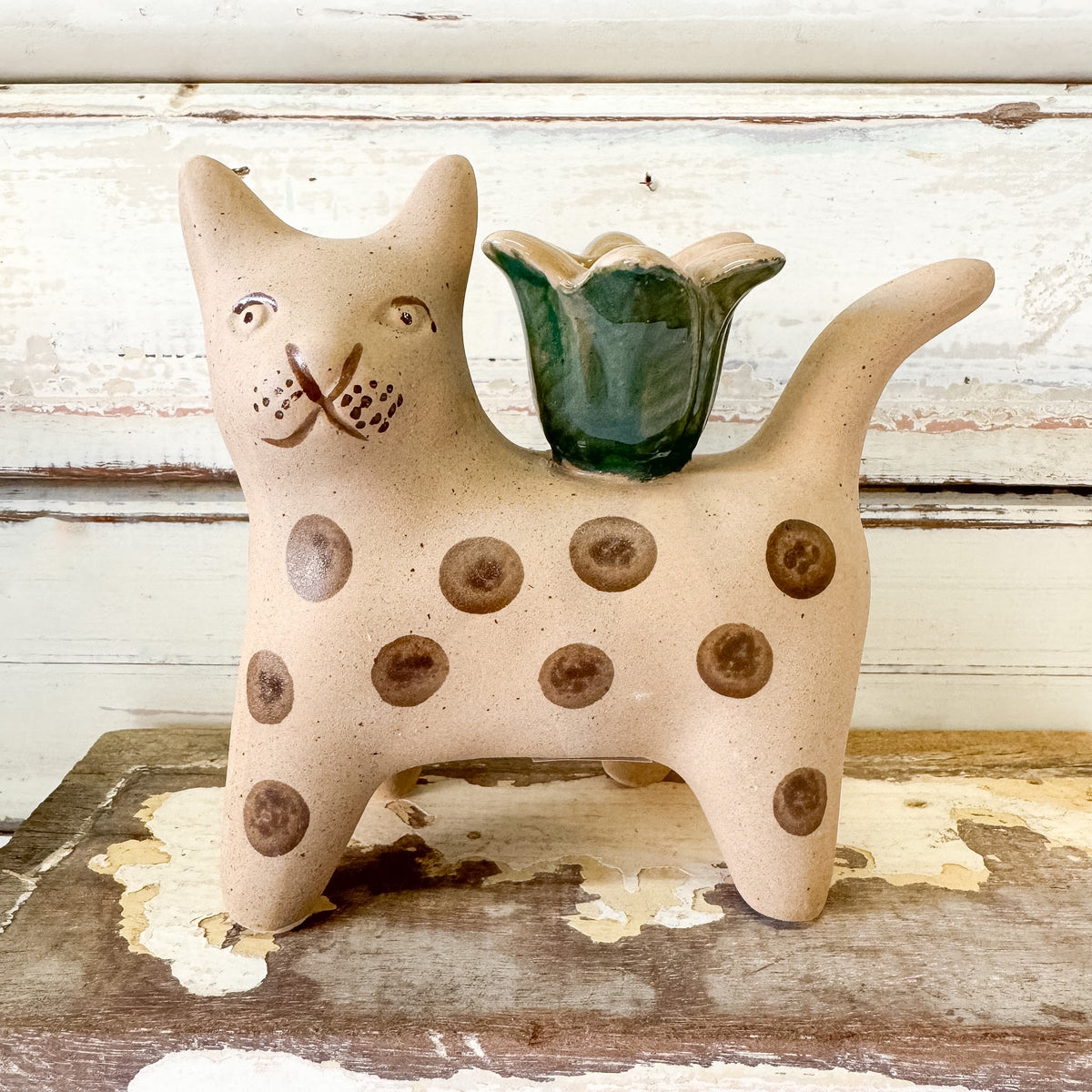 A ceramic cat-shaped candle holder or planter with a beige speckled body, brown spots, and a green flower-shaped top, displayed on a rustic wooden surface against a distressed white wood background.