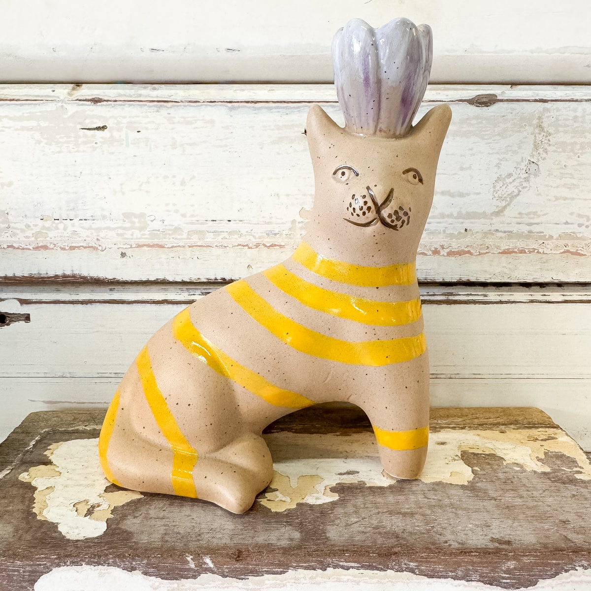 A ceramic cat figurine with a speckled beige body, yellow horizontal stripes, and a lavender-purple flower crown, sitting on a rustic wooden surface against a distressed white wood background.