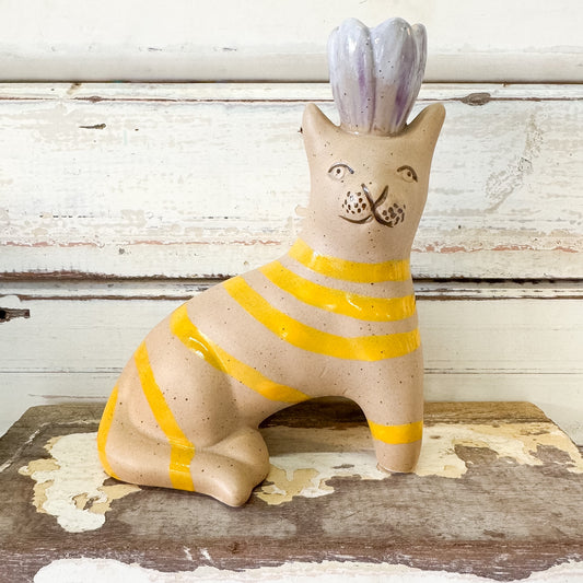 A ceramic cat figurine with a speckled beige body, yellow horizontal stripes, and a lavender-purple flower crown, sitting on a rustic wooden surface against a distressed white wood background.