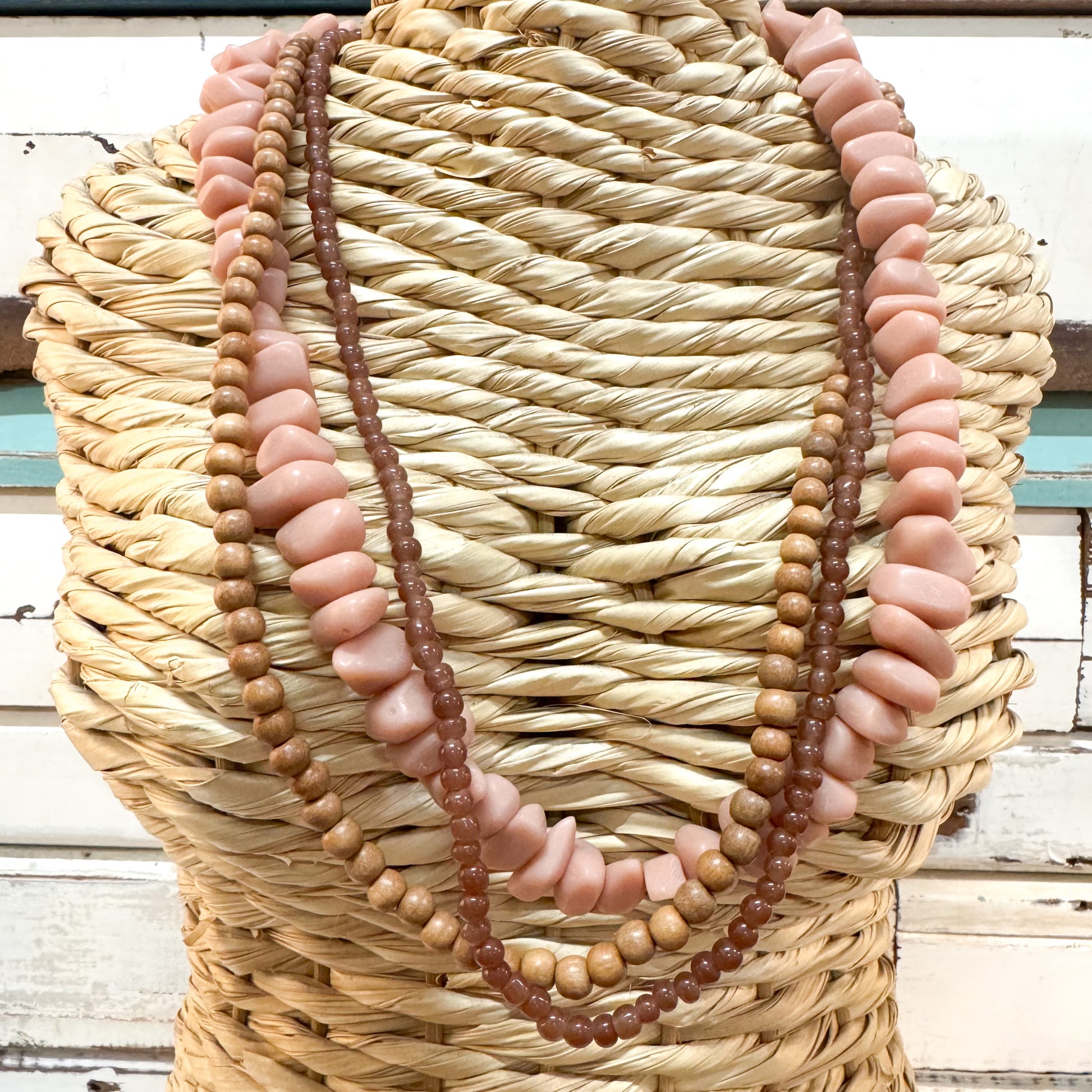 The Brightside Store | Sundaze Necklace ~ Coffee Break A multi-strand necklace displayed on a woven straw mannequin torso. The necklace features alternating strands of smooth, chunky dusty pink beads and small, polished brown wooden beads.