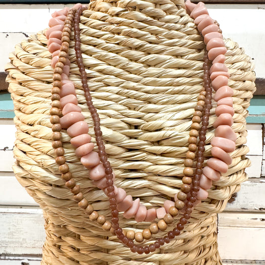 The Brightside Store | Sundaze Necklace ~ Coffee Break A multi-strand necklace displayed on a woven straw mannequin torso. The necklace features alternating strands of smooth, chunky dusty pink beads and small, polished brown wooden beads.