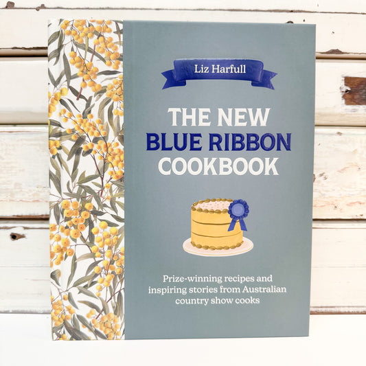 The New Blue Ribbon Cookbook by Liz Harfull, featuring a muted blue-gray cover with a yellow floral pattern on the spine and a small cake illustration with a blue ribbon, set against a rustic white wooden background.