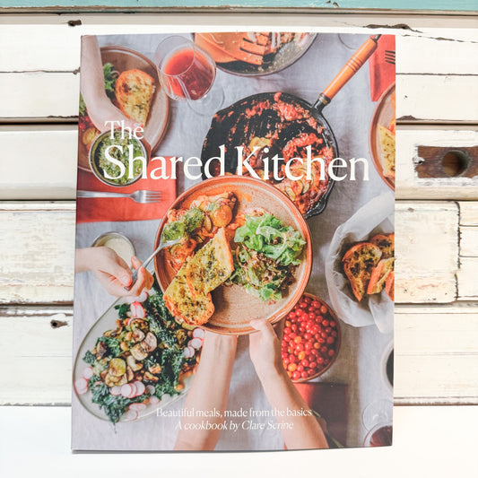 The Shared Kitchen cookbook by Clare Scribe, featuring a colorful overhead photograph of a shared meal on a rustic white wooden background.