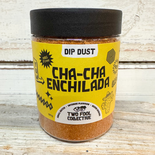 A jar of Two Fool Collective Cha-Cha Enchilada seasoning dip, featuring a bright yellow label and a black lid, set against a rustic white-painted wooden background.