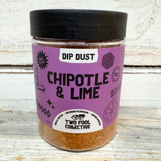 A clear plastic jar of Two Fool Collective Dip Dust in the Chipotle & Lime flavor, featuring a black lid and a purple label with black text, set against a rustic white wooden background.