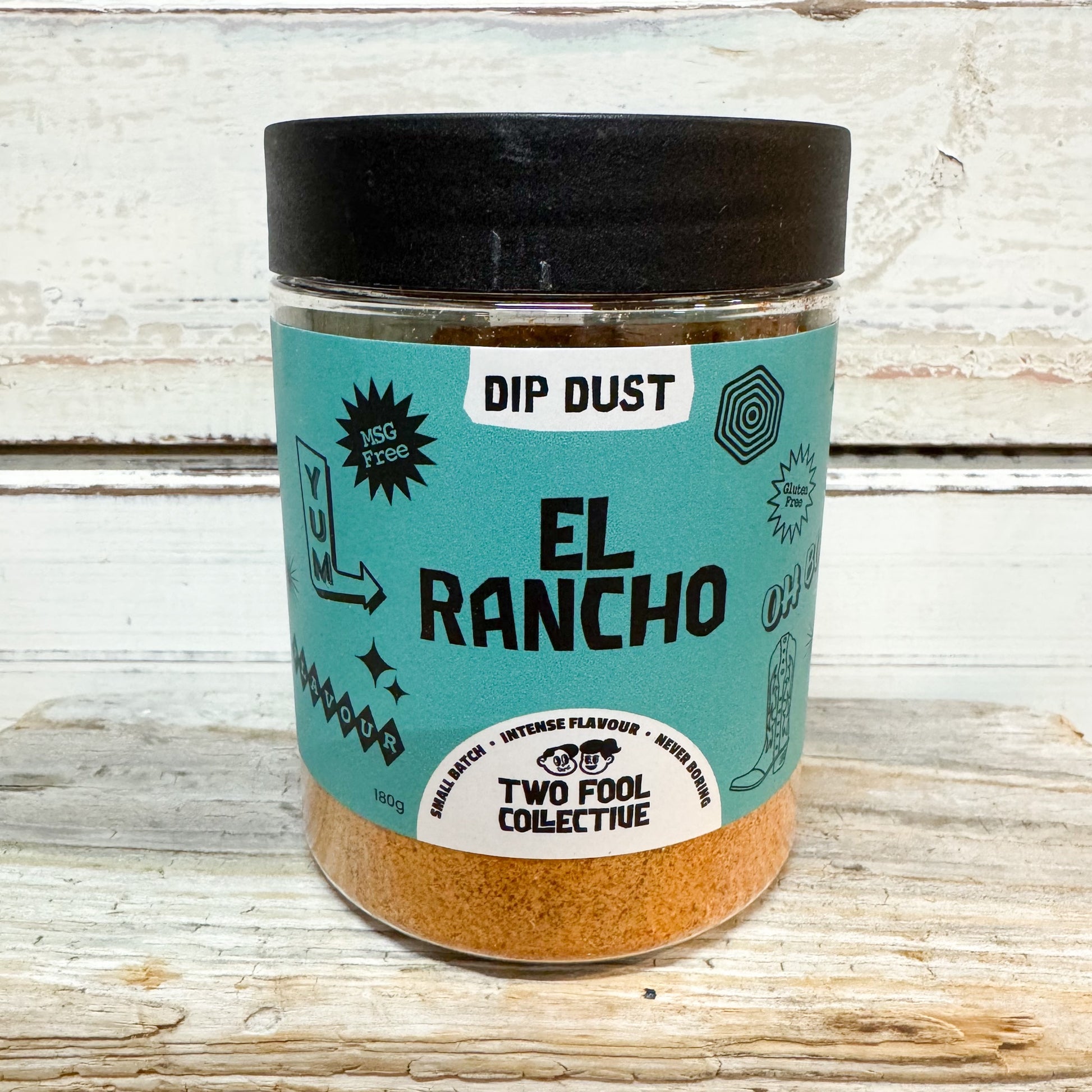 A clear plastic jar of El Rancho Dip Dust seasoning by Two Fool Collective, featuring a turquoise label and a black lid, set against a rustic white-washed wooden background.