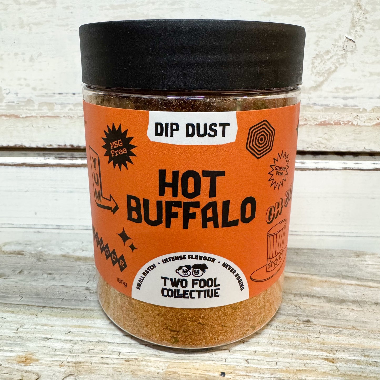 A clear plastic jar of Two Fool Collective 'Hot Buffalo' Dip Dust seasoning, featuring a bright orange label and a black lid, set against a rustic white-painted wooden background.