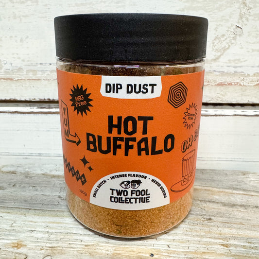 A clear plastic jar of Two Fool Collective 'Hot Buffalo' Dip Dust seasoning, featuring a bright orange label and a black lid, set against a rustic white-painted wooden background.