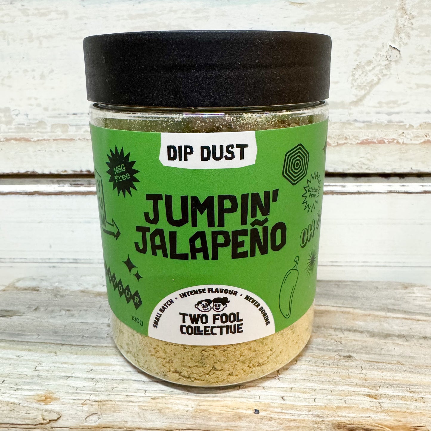 A clear plastic jar of Two Fool Collective 'Jumpin' Jalapeño' Dip Dust seasoning, featuring a bright green label and a black lid, set on a rustic white wooden surface.