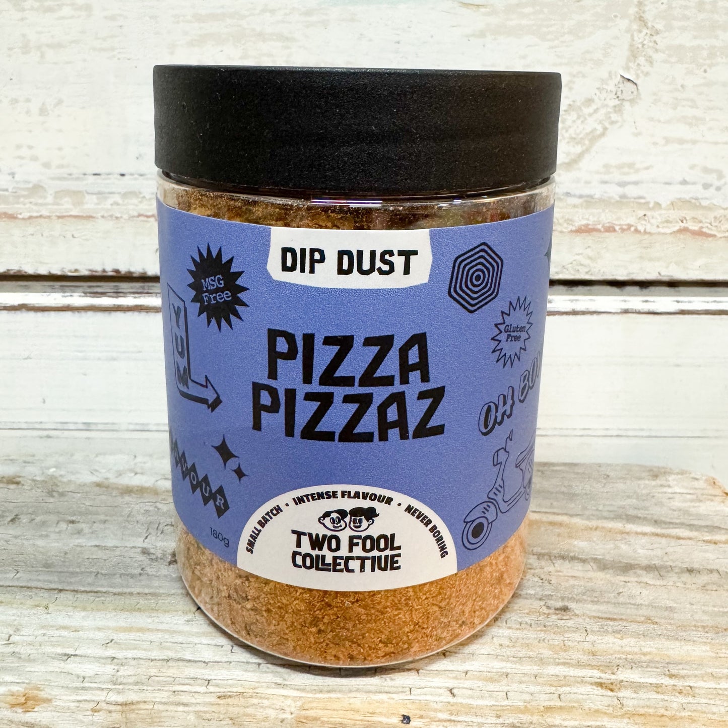 A clear plastic jar of Two Fool Collective Dip Dust Pizza Pizzaz seasoning, featuring a black lid and a light blue label, set against a rustic white-washed wooden background.