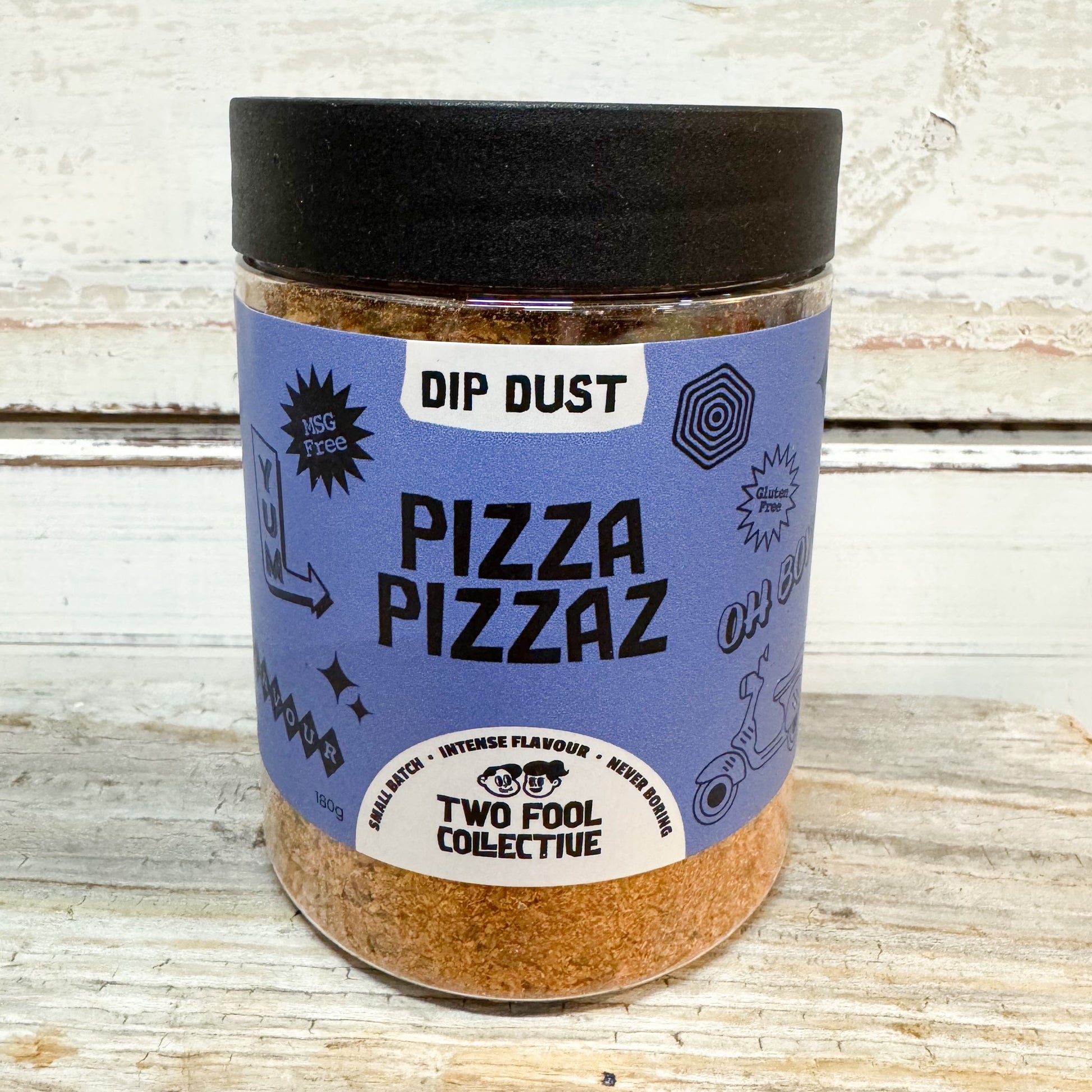 A clear plastic jar of Two Fool Collective Dip Dust Pizza Pizzaz seasoning, featuring a black lid and a light blue label, set against a rustic white-washed wooden background.