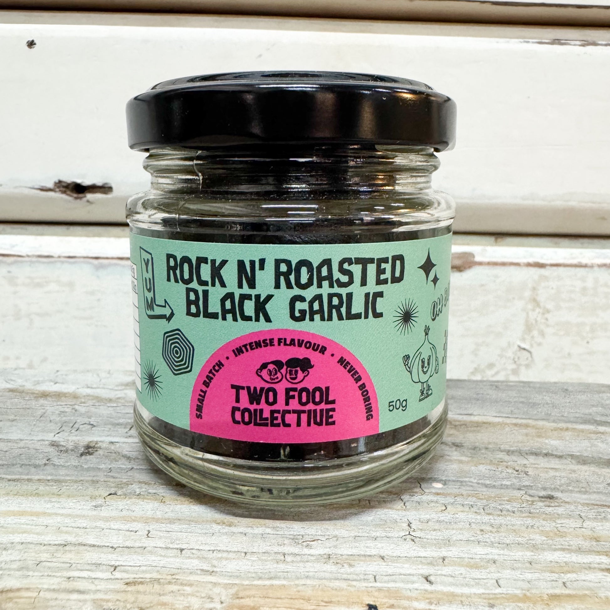A glass jar of Two Fool Collective Rock N' Roasted Black Garlic with a black lid and a mint green and pink label, set on a rustic white wooden surface.
