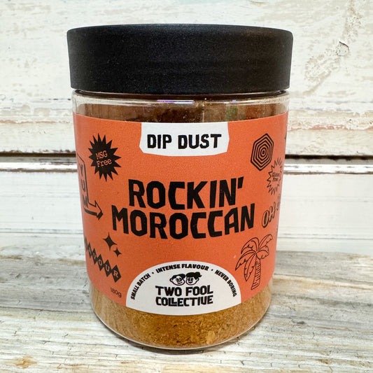 A clear plastic jar of Two Fool Collective 'Rockin' Moroccan' Dip Dust seasoning with a black lid and an orange label, set on a rustic white wooden surface.
