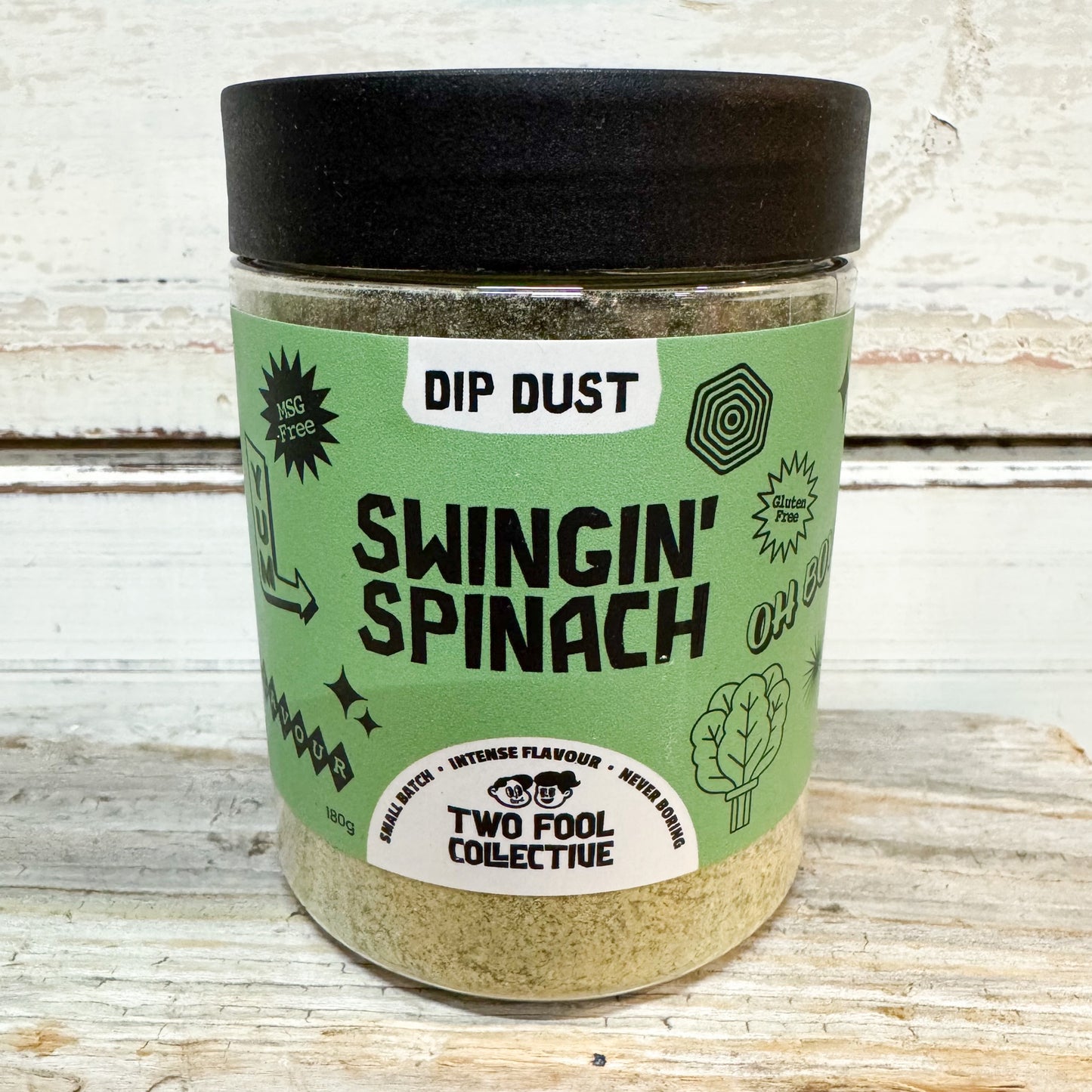 A clear plastic jar of Two Fool Collective Dip Dust seasoning, featuring a light green label with black text and a black lid, set against a rustic white wooden background.