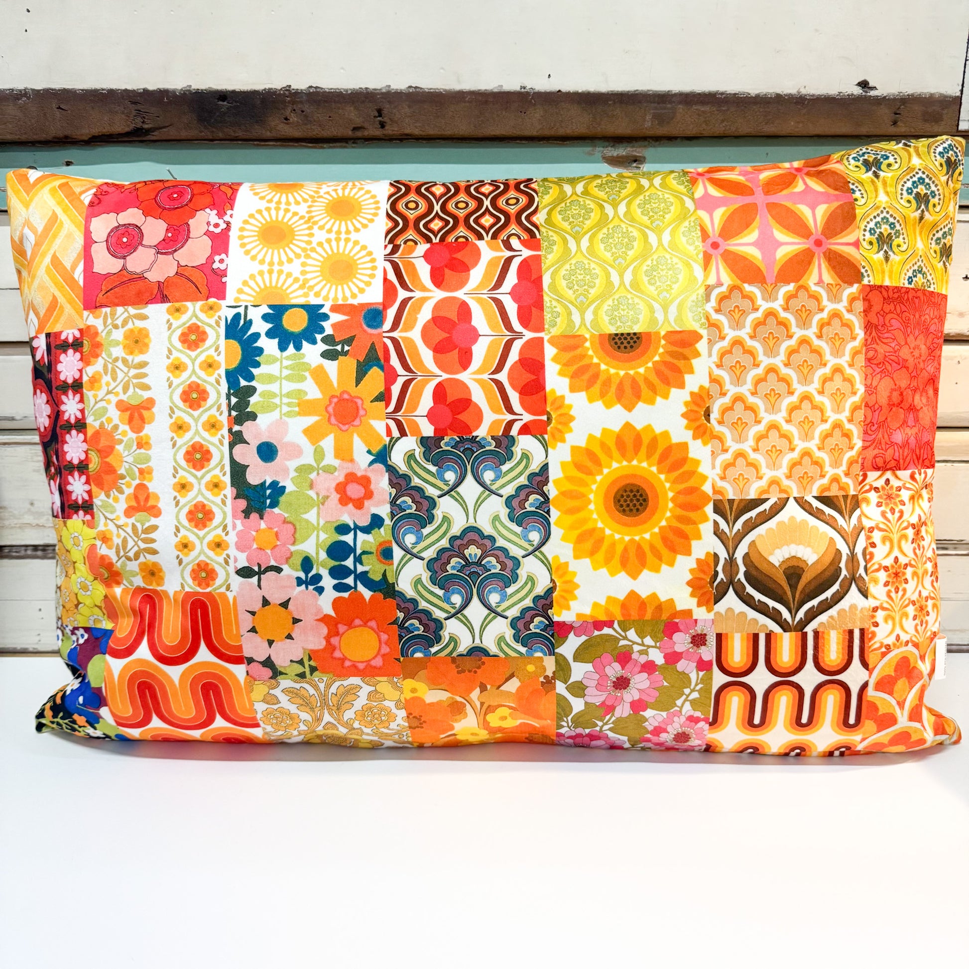A square patchwork pillow cover featuring a vibrant retro 1970s and 1980s floral, geometric, and sunflower pattern in orange, yellow, red, blue, and green, displayed on a white wooden surface.