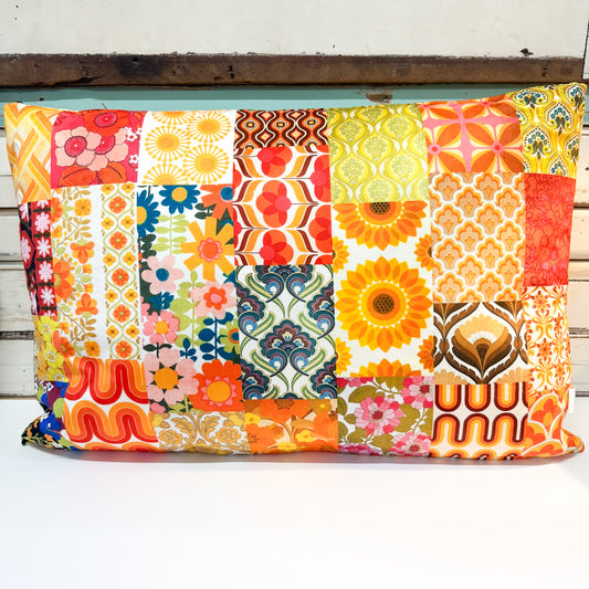 A square patchwork pillow cover featuring a vibrant retro 1970s and 1980s floral, geometric, and sunflower pattern in orange, yellow, red, blue, and green, displayed on a white wooden surface.