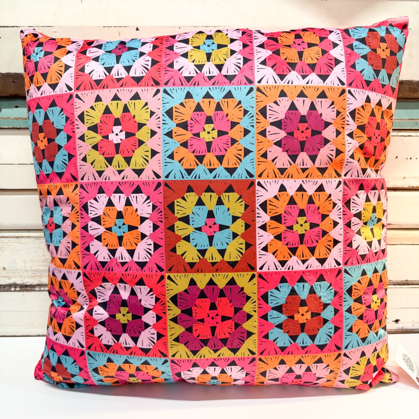 A square throw pillow featuring a vibrant, multicolor retro granny square patchwork pattern, set against a rustic white wooden plank background.