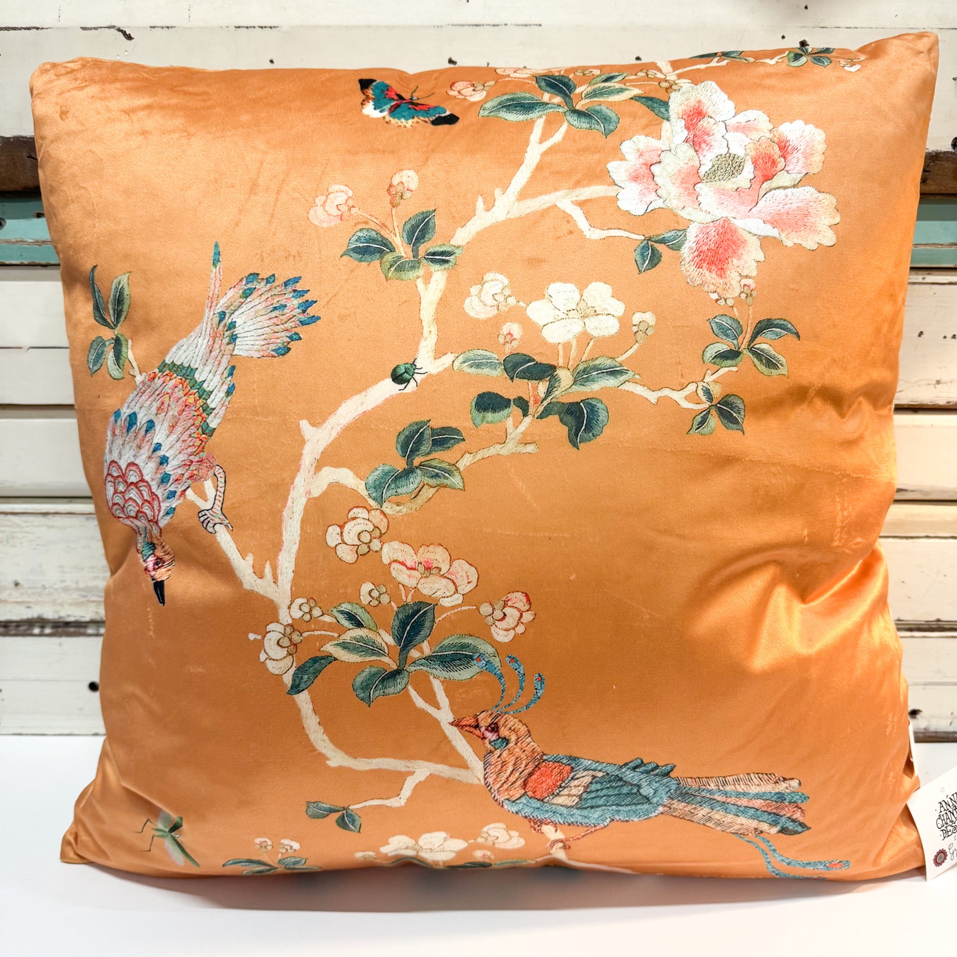 An orange satin throw pillow featuring a traditional Chinese Chinoiserie print of birds, peonies, and branches, displayed against a rustic white wooden background.