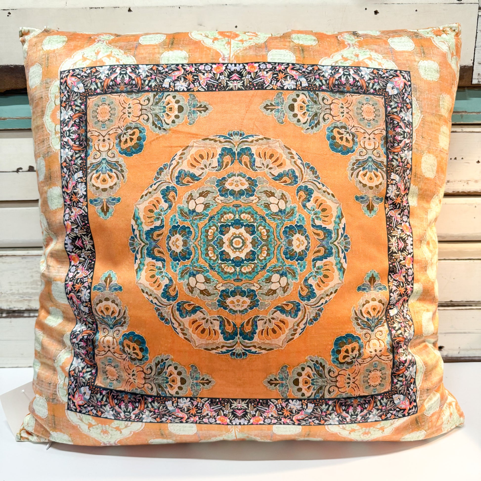 A square decorative throw pillow featuring an intricate traditional Persian-style floral pattern in orange, teal, beige, and dark navy, set against a rustic white wooden background.