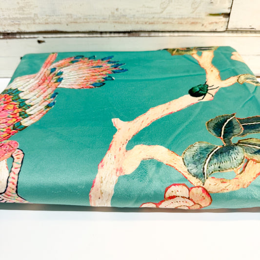 A folded piece of teal silk fabric featuring a traditional Chinese-style print with a phoenix, branch, leaves, and a beetle, displayed on a white wooden surface.
