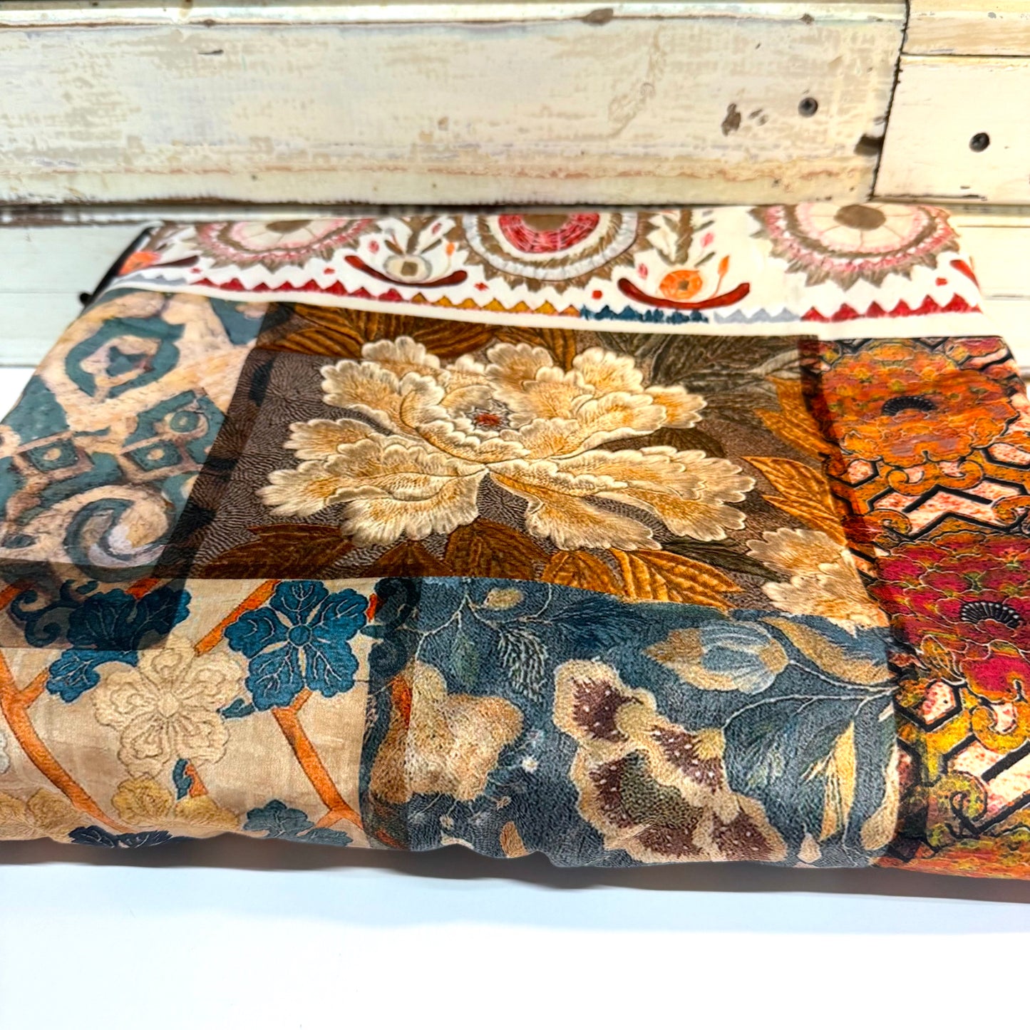 A folded patchwork fabric featuring a variety of vintage floral and paisley patterns in muted tones of blue, beige, brown, orange, and red, displayed against a rustic white wooden background.