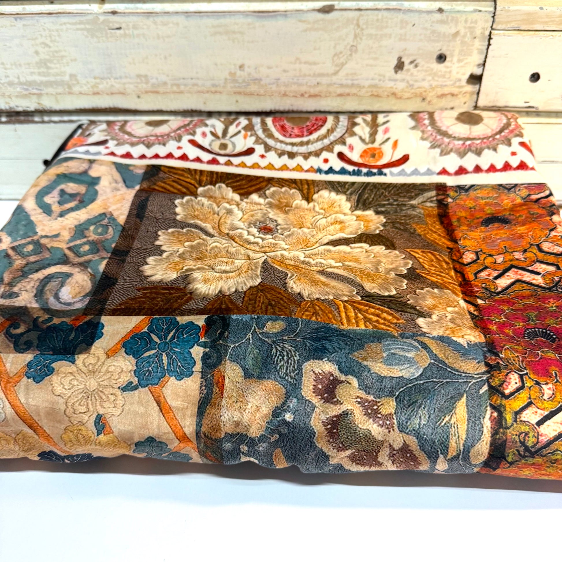 A folded patchwork fabric featuring a variety of vintage floral and paisley patterns in muted tones of blue, beige, brown, orange, and red, displayed against a rustic white wooden background.