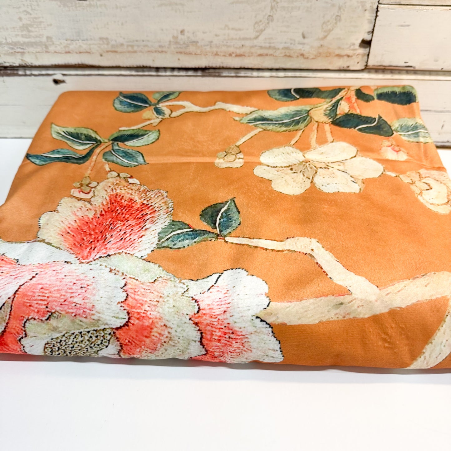 A folded piece of orange fabric with a detailed floral print featuring white blossoms, pink and red petals, and green leaves, displayed on a white surface.