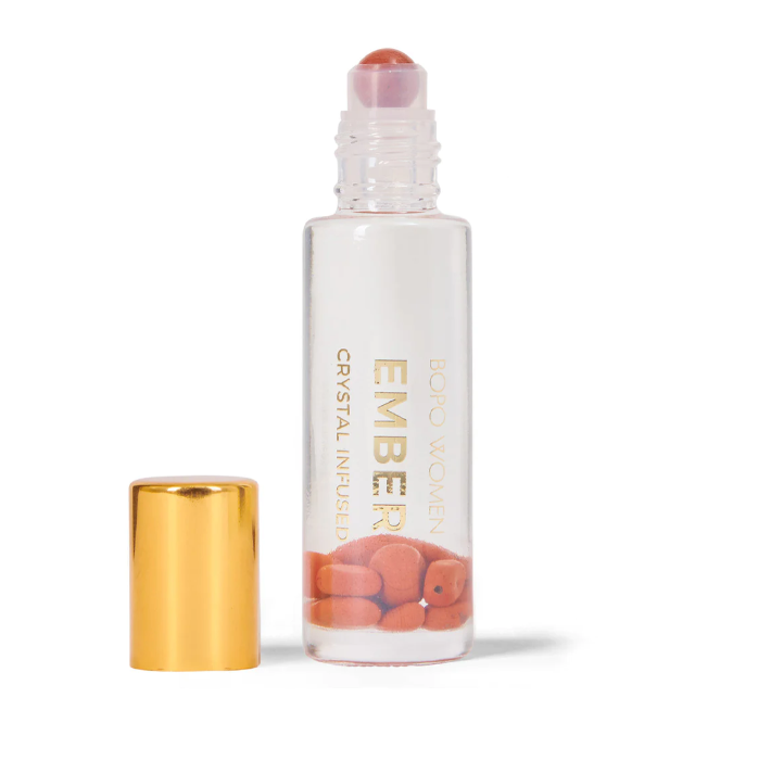 A clear rollerball perfume dispenser with a gold-colored cap and a red jasper roller beneath it. The label reads 'EMBER WOMEN crystal infused essential oil blend'.