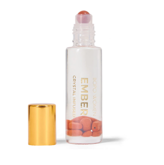 A clear rollerball perfume dispenser with a gold-colored cap and a red jasper roller beneath it. The label reads 'EMBER WOMEN crystal infused essential oil blend'.