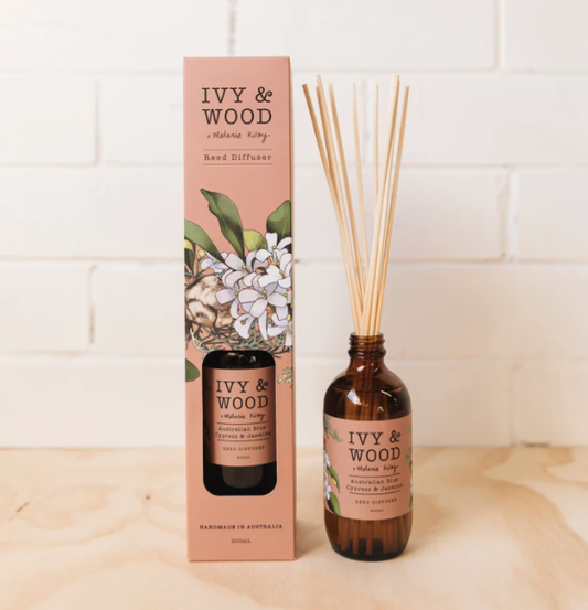 An Ivy & Wood reed diffuser with Australian Blue Cypress & Jasmine fragrance, displayed next to its packaging box, on a white surface against a white wall, with rattan reeds hanging from the bottle.