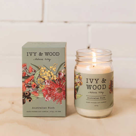 Australian Bush Candle by Ivy & Wood in a clear glass jar with a burning candle inside, alongside its packaging box featuring floral illustrations.