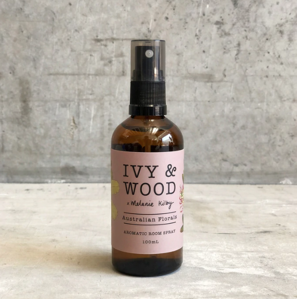 A brown glass bottle with a spray nozzle, labeled 'IVY & WOOD Australian Florals Aromatic Room Spray 100ml'. The label features floral illustrations and the product is positioned against a concrete background.
