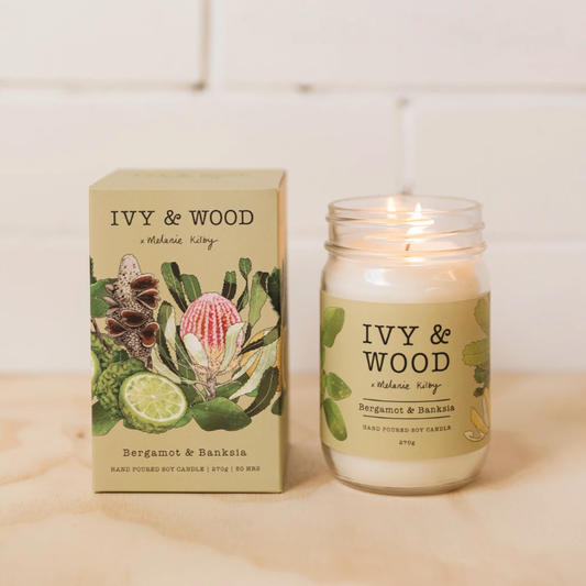 A Ivy & Wood Bergamot & Banksia candle in a white jar with a label featuring an illustration of native Australian flora. The product is displayed with its packaging, which is also designed with native Australian flora.