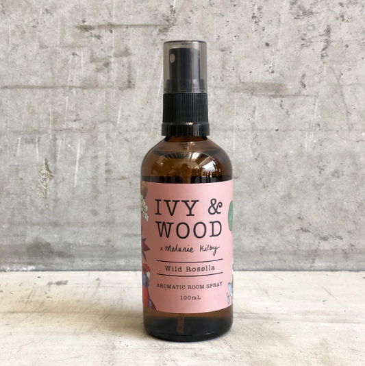 A bottle of Ivy & Wood Wild Rosella aromatic room spray with a pink label and green leaves illustration, on a concrete background, 100ml volume indicated.