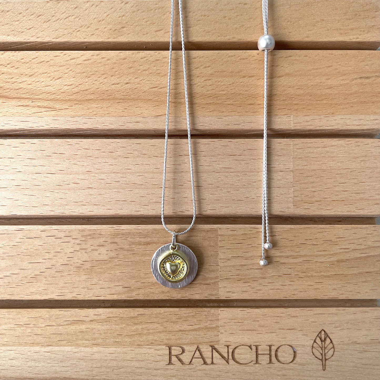 RANCHO DESIGN Full Heart Gold Necklace A matte gold necklace with a textured disc pendant featuring a heart design, attached to a silver chain with a bead.
